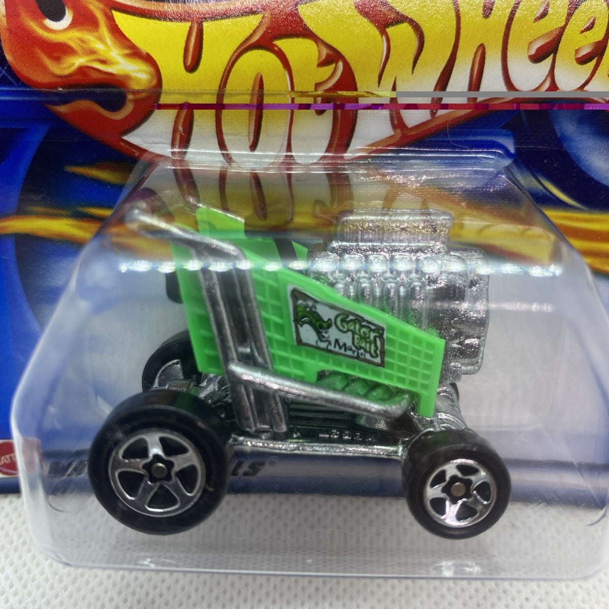 Green truck toy with black chassis in clear blister, Hot Wheels flames design behind